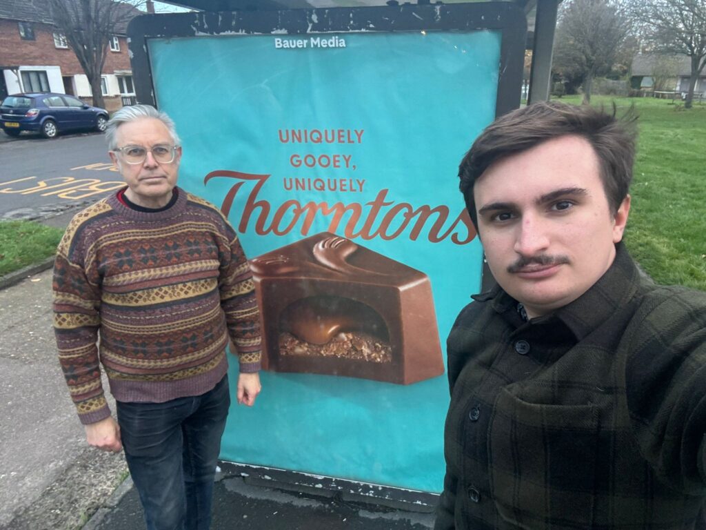 Image: Green County Councillors Darren Green (Romsey) and Elliot Tong (Abbey) in front of a bus shelter advertisement in Cambridge.