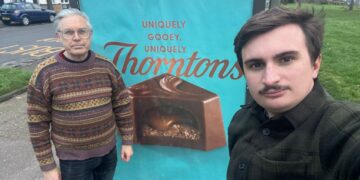 Image: Green County Councillors Darren Green (Romsey) and Elliot Tong (Abbey) in front of a bus shelter advertisement in Cambridge.